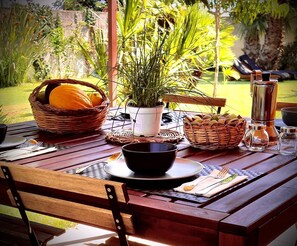 Una mesa de madera al aire libre con una cesta de fruta, una planta en maceta y un molinillo de café.
