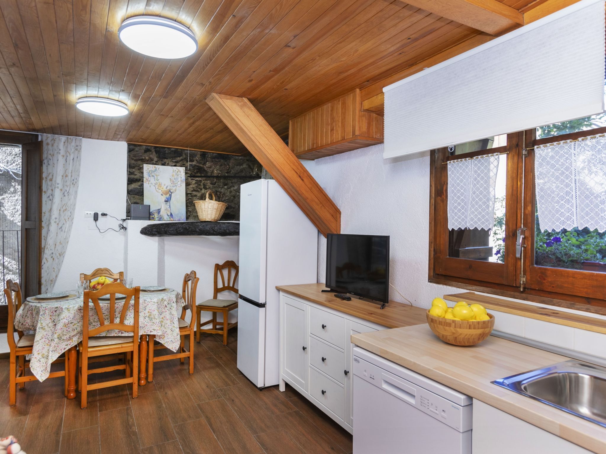 A kitchen with wooden cabinets, a white refrigerator, a dining table with chairs, a television, and a window with lace curtains.
