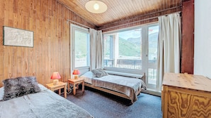 Habitación con paneles de madera, dos camas, una mesita con lámpara y ventanas amplias con vistas al exterior.