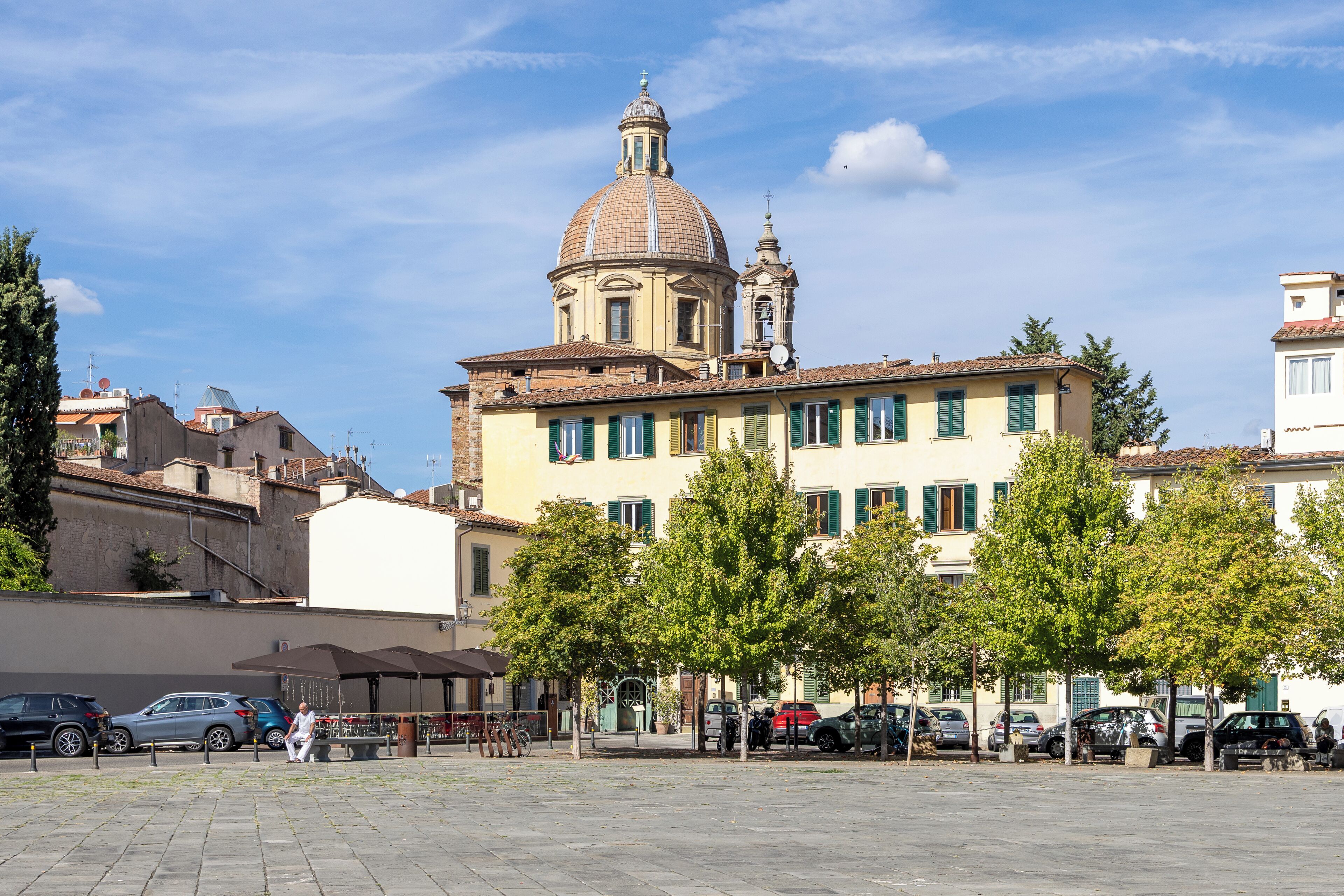 A historic building with a prominent dome and bell tower, surrounded by parked cars and trees.