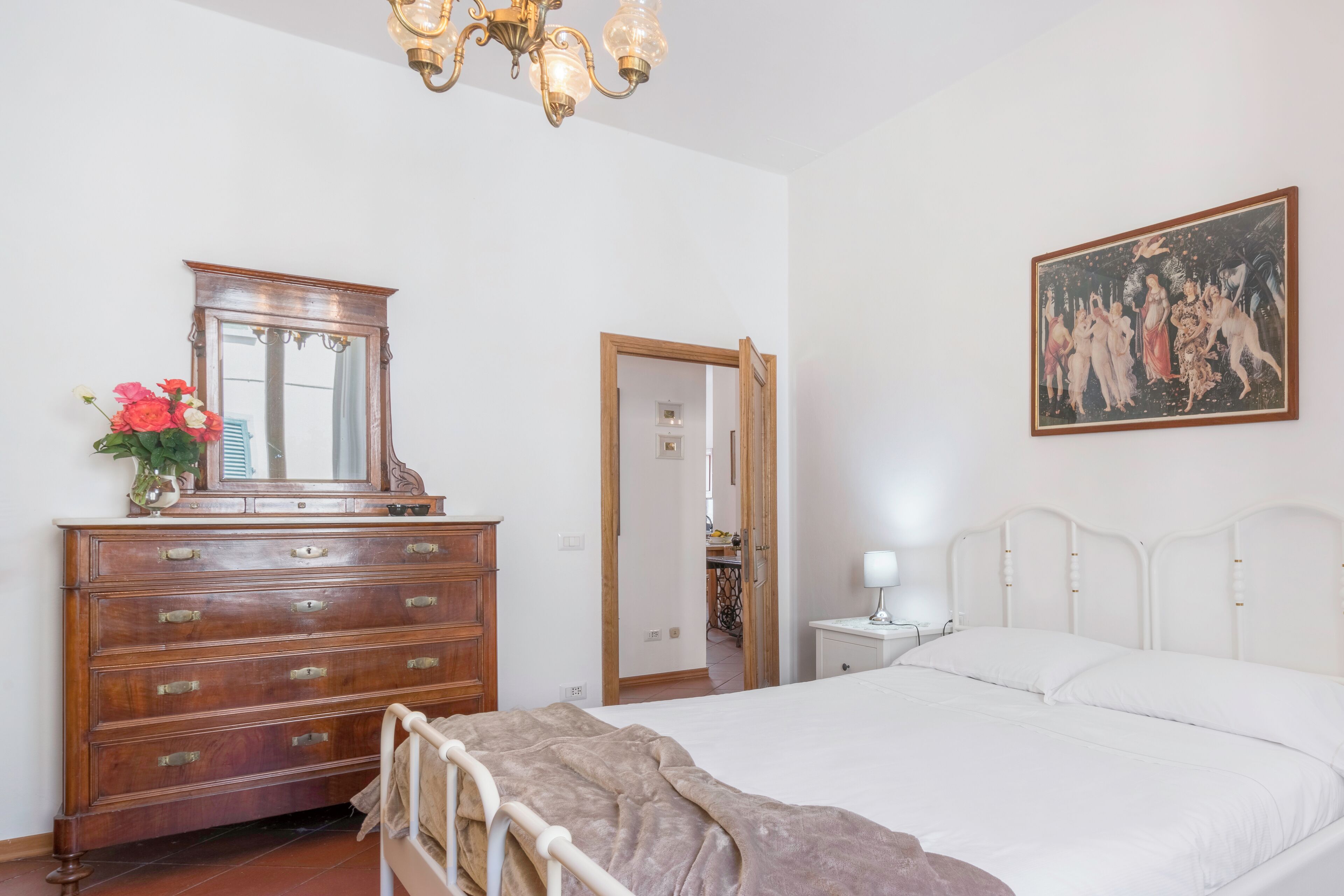 Ein Schlafzimmer mit einem hölzernen Schminktisch, einem Bett mit weißer Bettwäsche, einer Chandelieber, einem Bild an der Wand und einem Spiegel.