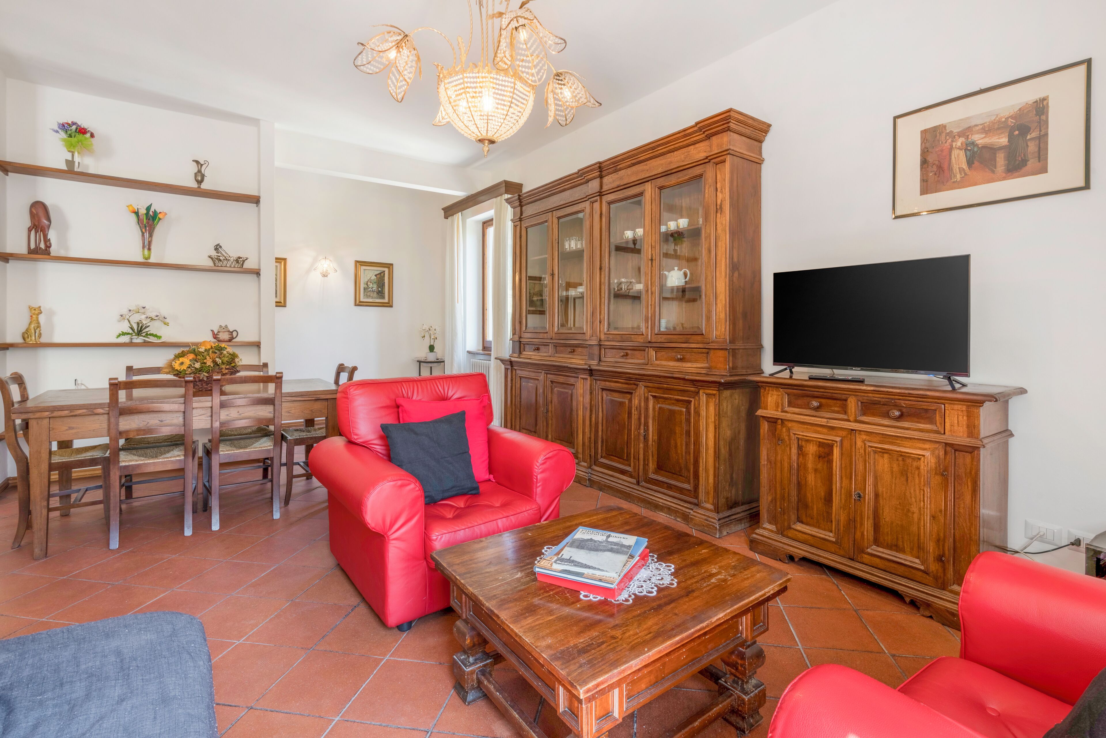 Een woonkamer met een rode bank, een houten eettafel met stoelen, een rode fauteuil, een houten kast en een flatscreen televisie.