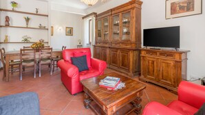 A living room with a red sofa, wooden dining table, and chairs, a red armchair, a wooden cabinet, and a flat-screen TV. - Chiara Apartment In Porta San Frediano (Florence)