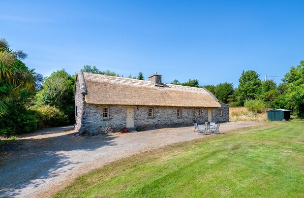 A stone house with a thatched roof, a gravel driveway, and a small outdoor seating area. - Traditional style thatched cottage in Kenmare (Kenmare)