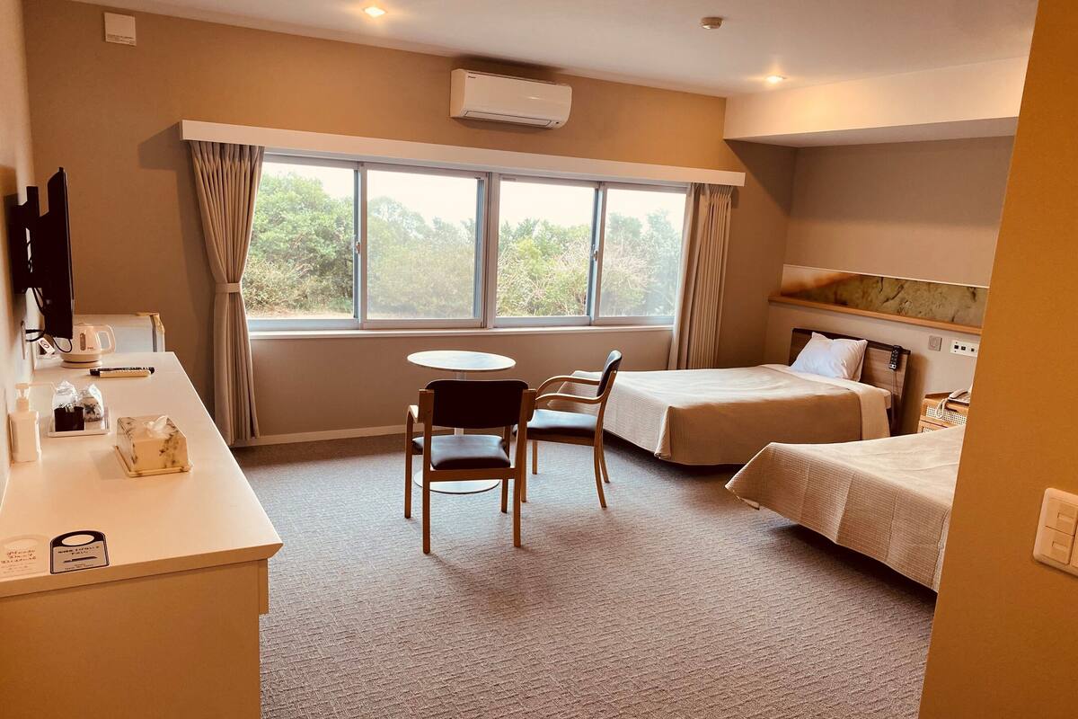A hotel room with two beds, a desk with a chair, a small table, and a window with curtains.