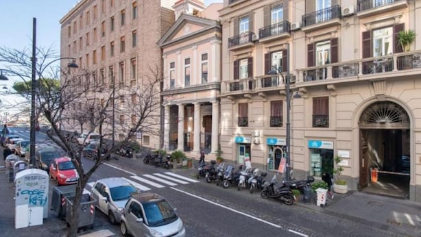 Vista de la calle con una hilera de edificios de varios pisos, coches aparcados y motocicletas.