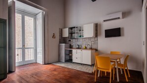 Een compacte keuken met witte kastjes, een kleine koelkast, en een eethoek met een houten tafel en gele stoelen.