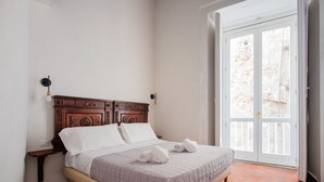 Een slaapkamer met een houten hoofdbord, een bed met witte lakens, een nachtkastje en een groot raam met uitzicht op een stenen muur.
