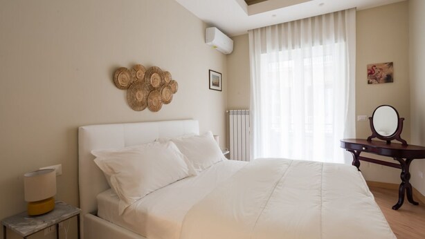A bedroom with a large bed, a bedside table, a wall-mounted air conditioner, a radiator, a window with sheer curtains, and a small table with a mirror. - Da Luca e Laura by Dimorra (Napoli)