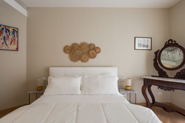 A neatly made bed with white linens, a decorative wall hanging, a framed picture, and a wooden dresser with a mirror.