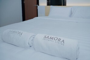 A neatly made bed with white linens and two rolled towels with the text "SAMORA" and "PLACEFUL LIVING". - The Samora Living (Ngaglik)