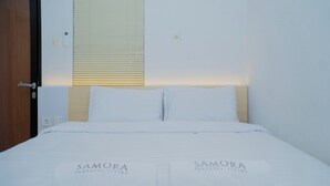 Una cama bien hecha con ropa blanca y dos toallas con el texto "SAMORA PEACEFUL LIVING", una puerta de madera, un interruptor de luz y una ventana con persianas.