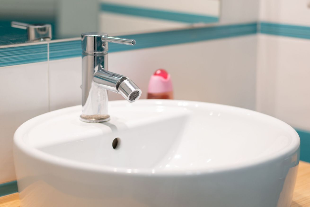 Ein weißes Waschbecken mit einem chromfarbenen Wasserhahn und einer kleinen Seifenflasche im Hintergrund.