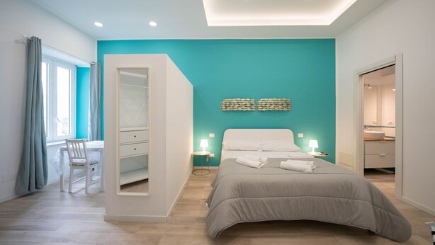 A modern bedroom with a teal accent wall, a bed with a grey duvet, a desk with a chair, and a walk-in closet.