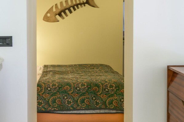 A room with a bed covered in a patterned bedspread, a wall-mounted light fixture, and a wooden dresser. - Orabon Suite By Dimorra (Napoli)
