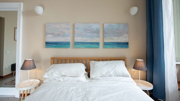 Habitación de hotel con cama de madera, ropa de cama blanca, dos mesitas de noche con lámparas y tres cuadros colgados en la pared que representan una playa.