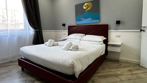 A neatly made bed with white linens and towels, a red headboard, and a painting of a wave on the wall. - Dimorra Boutique room 2 (Napoli)