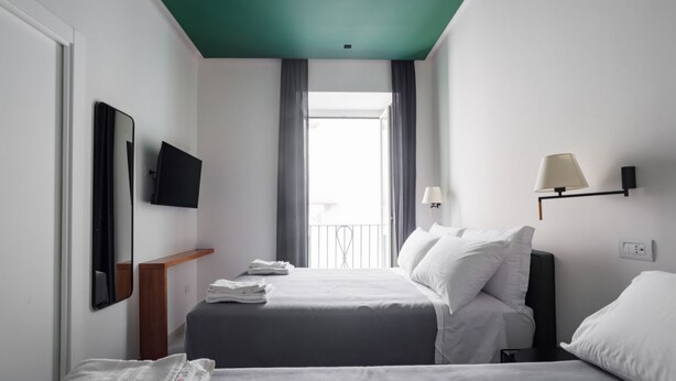 A hotel room with two beds, a TV mounted on the wall, a window with curtains, and a lamp on the nightstand. - Weaver Suites 2 By Dimorra (NAPOLI)