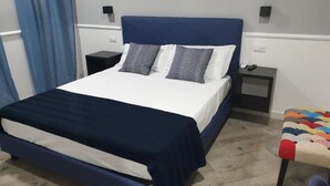 A bed with a blue headboard, white bedding, and two patterned pillows. There is a nightstand with a remote control and a small table with a colorful cushion.