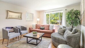 Ein Wohnzimmer mit einer Couch, zwei Sesseln, einem Couchtisch und einem großen Fenster mit Vorhängen.