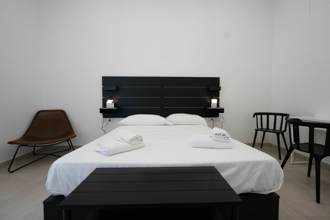 A neatly made bed with white linens, a black headboard, and bedside lamps. There is a brown chair and a small table with a chair.