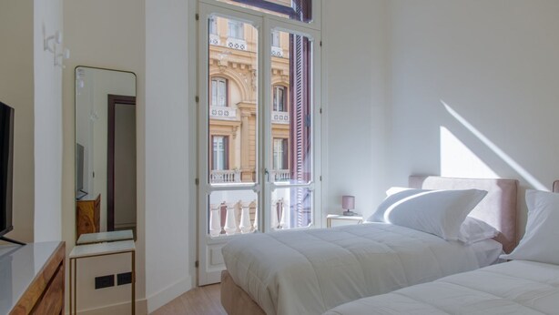 Un dormitorio con cama, mesita de noche, espejo y ventana con vistas a un edificio.