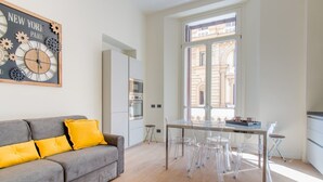 Una sala moderna con un sofá gris, sillas transparentes, una mesa de comedor de vidrio y un amplio ventanal con vistas a edificios.