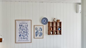 A living room with a white paneled wall, a framed poster of seashells, a small framed seashell artwork, and a wooden shelf with pottery.