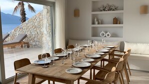 A dining area with a wooden table set for six, surrounded by wooden chairs, with a view of the sea and mountains through large windows.