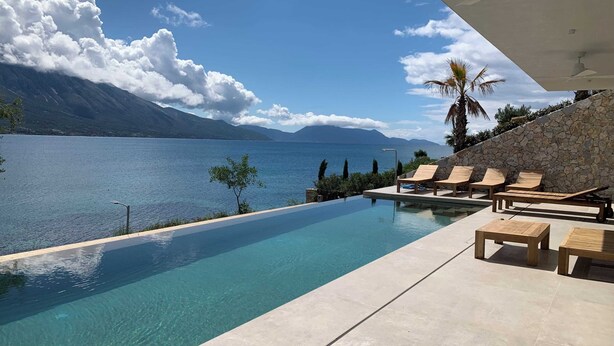 An infinity pool with a clear view of a lake and mountains, wooden lounge chairs, and a stone wall.