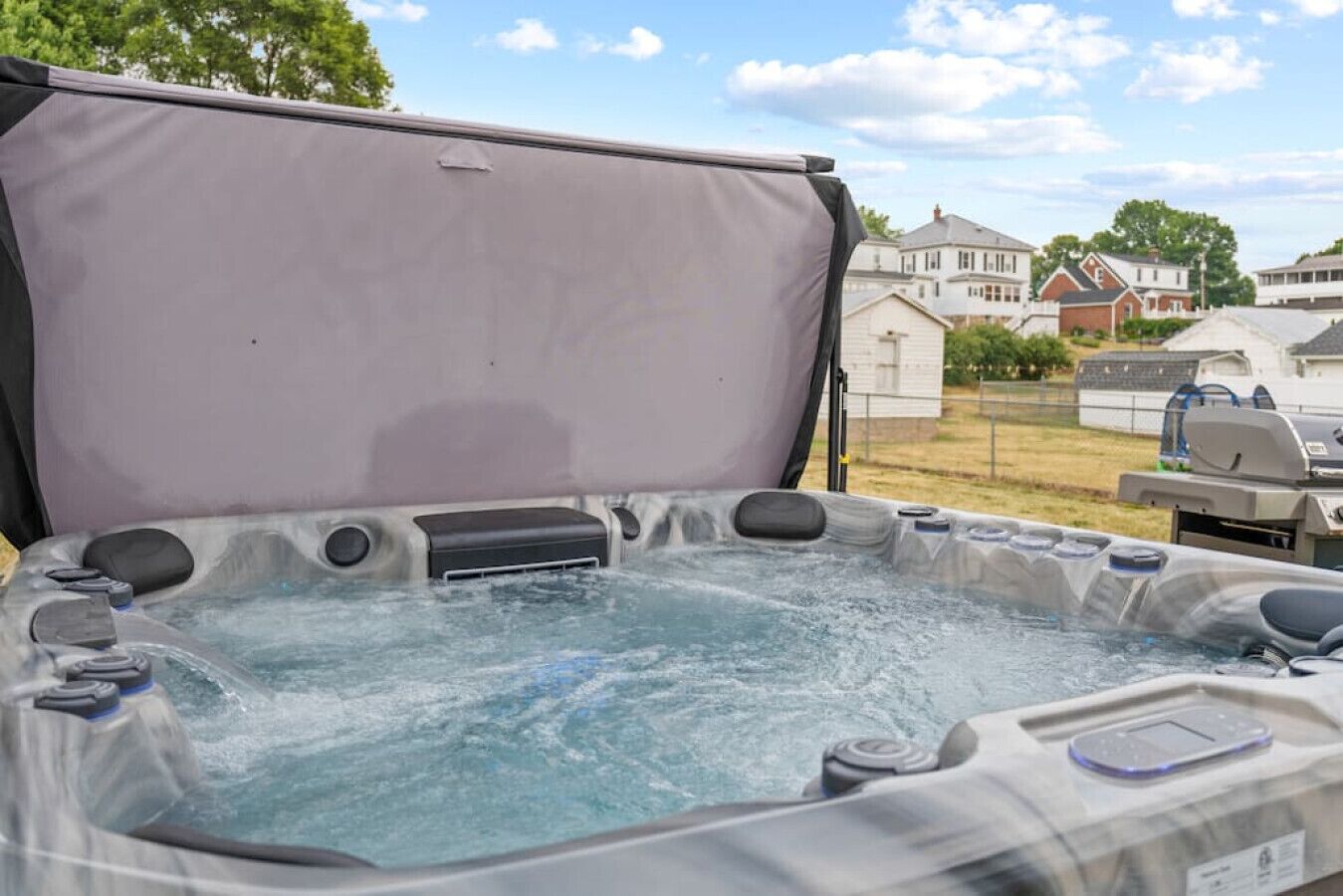 A hot tub with a cover, surrounded by a residential area with houses and a barbecue grill in the background.