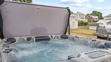 A hot tub with a cover, surrounded by a residential area with houses and a barbecue grill in the background.