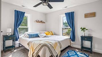 A bedroom with a bed, two nightstands, a ceiling fan, a window with curtains, a welcome sign, and a small plant.