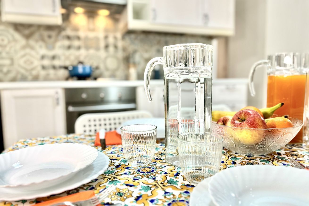 A dining table with a patterned tablecloth, a pitcher, a bowl of fruit, and clear glasses.