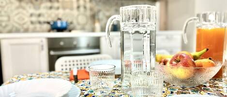 A dining table with a patterned tablecloth, a pitcher, a bowl of fruit, and clear glasses.