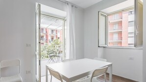 Una habitación luminosa con una mesa y sillas blancas, un balcón con vistas a un edificio y una ventana con vistas a un edificio.