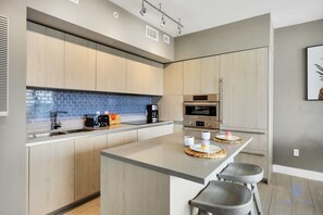 A modern kitchen with light wood cabinets, a central island with stools, a built-in oven, and a coffee machine.