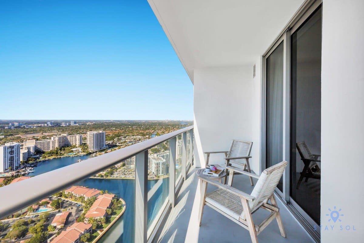 A balcony with a view of a river and buildings, furnished with two chairs and a small table.