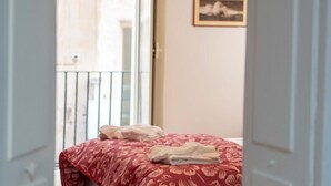 Une chambre à coucher avec un lit recouvert d’une couvre-lit à motifs floraux, une fenêtre donnant sur un immeuble et un tableau encadré accroché au mur.
