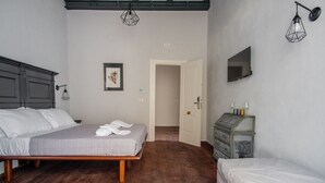 Een slaapkamer met een houten vloer, een bed met een grijze plaid, een dressoir en een televisie aan de muur.