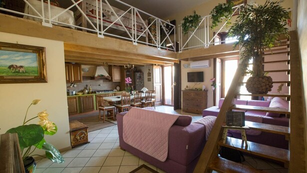 A spacious loft-style living area with a high ceiling, a purple sofa, a dining table, and a kitchen in the background. - Suite Orchidea by Dimorra (Napoli)