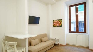 A living room with a beige sofa, a white table, a TV mounted on the wall, a colorful painting, and a window with a view. - Neapolitan Soul 1 By Dimorra (Napoli)