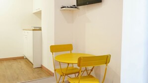 Een kleine keuken met een witte koelkast, een magnetron en een spoelbak. Er staat een gele tafel met twee stoelen.