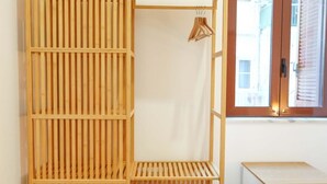 Een kamer met een houten kledingkast met legplanken, een klein tafeltje en een raam met jaloezieën.