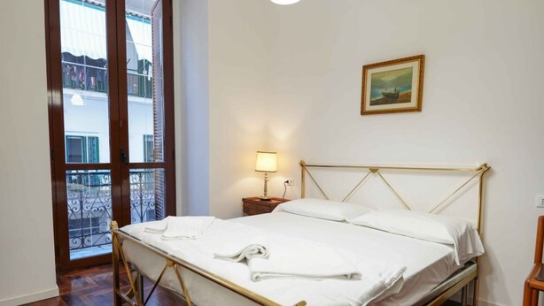 A neatly made bed with white linens and a towel, a bedside lamp, and a painting on the wall. - Neapolitan Soul 3 By Dimorra (Napoli)