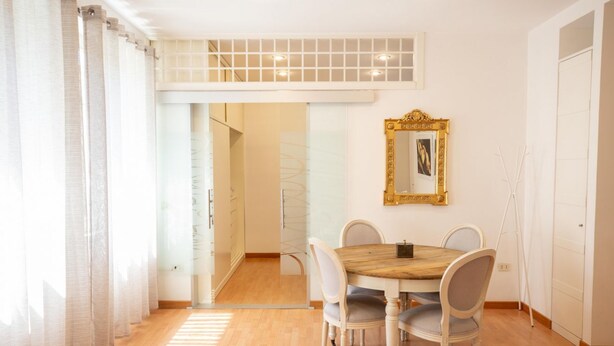 A dining area with a wooden table and white chairs, a gold-framed mirror on the wall, and a view of a glass door leading to a balcony. - Star in Toledo By Dimorra (Napoli)