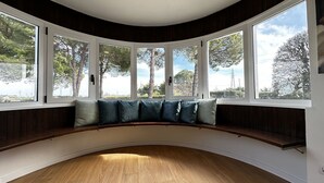 Ein Zimmer mit einer gebogenen Holzbank unter großen Fenstern mit weißen Rahmen, mit Blick auf Bäume und einen klaren Himmel.