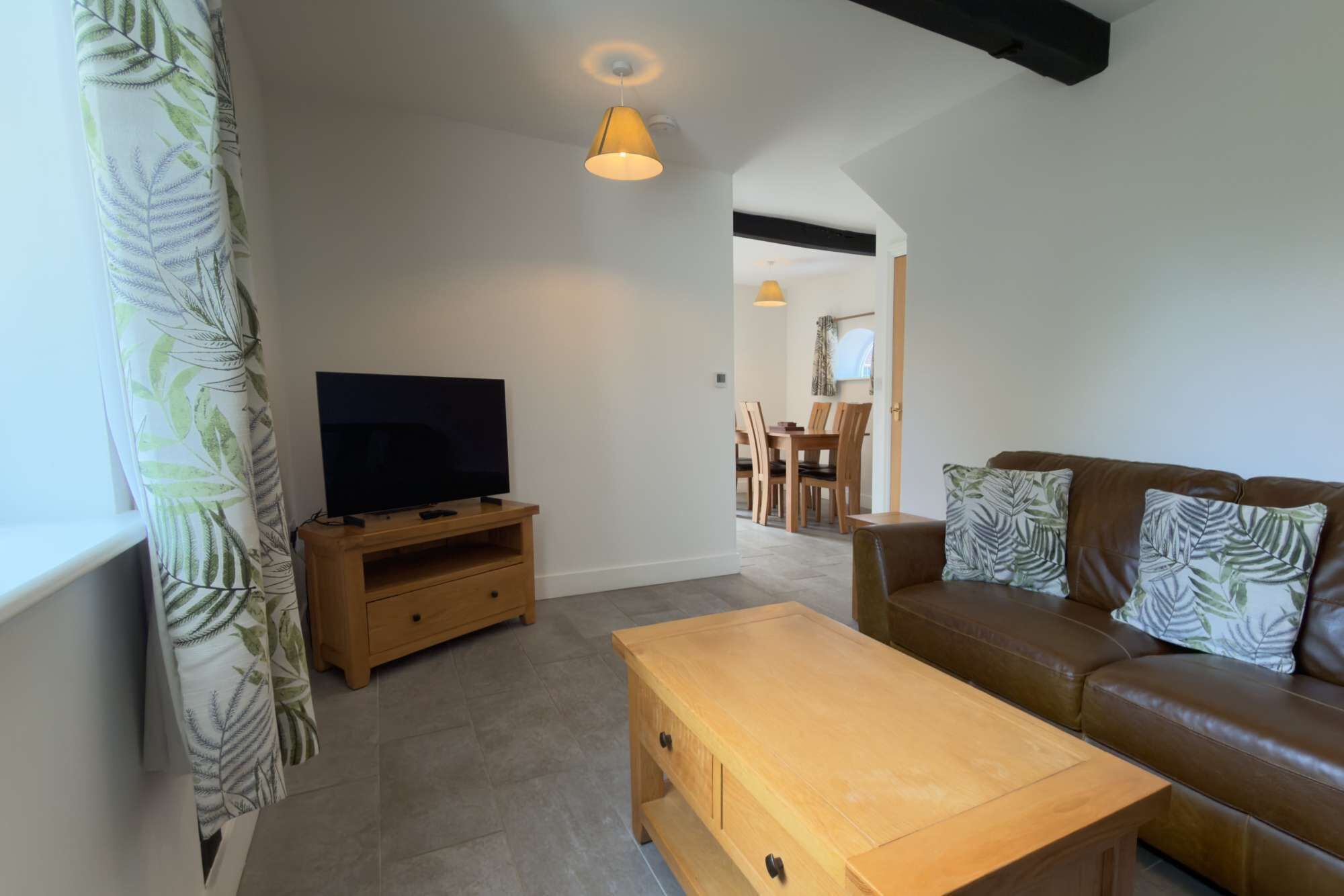 Ein Wohnzimmer mit Fernseher auf einem Holzboden, ein braunes Ledersofa mit gemusterten Kissen, ein Holztisch und ein Essbereich im Hintergrund.
