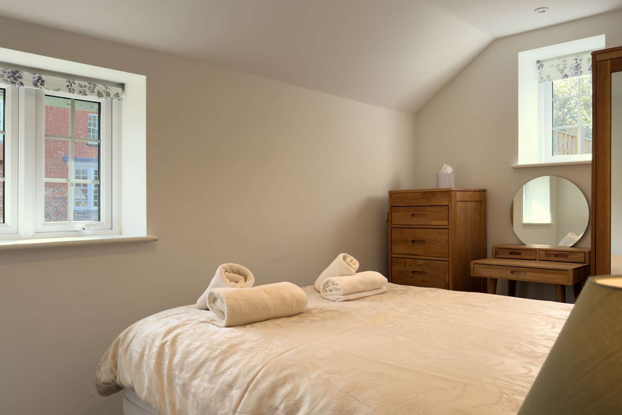 A bedroom with a bed, a wooden dresser, a mirror, and a window with a view of a brick building.