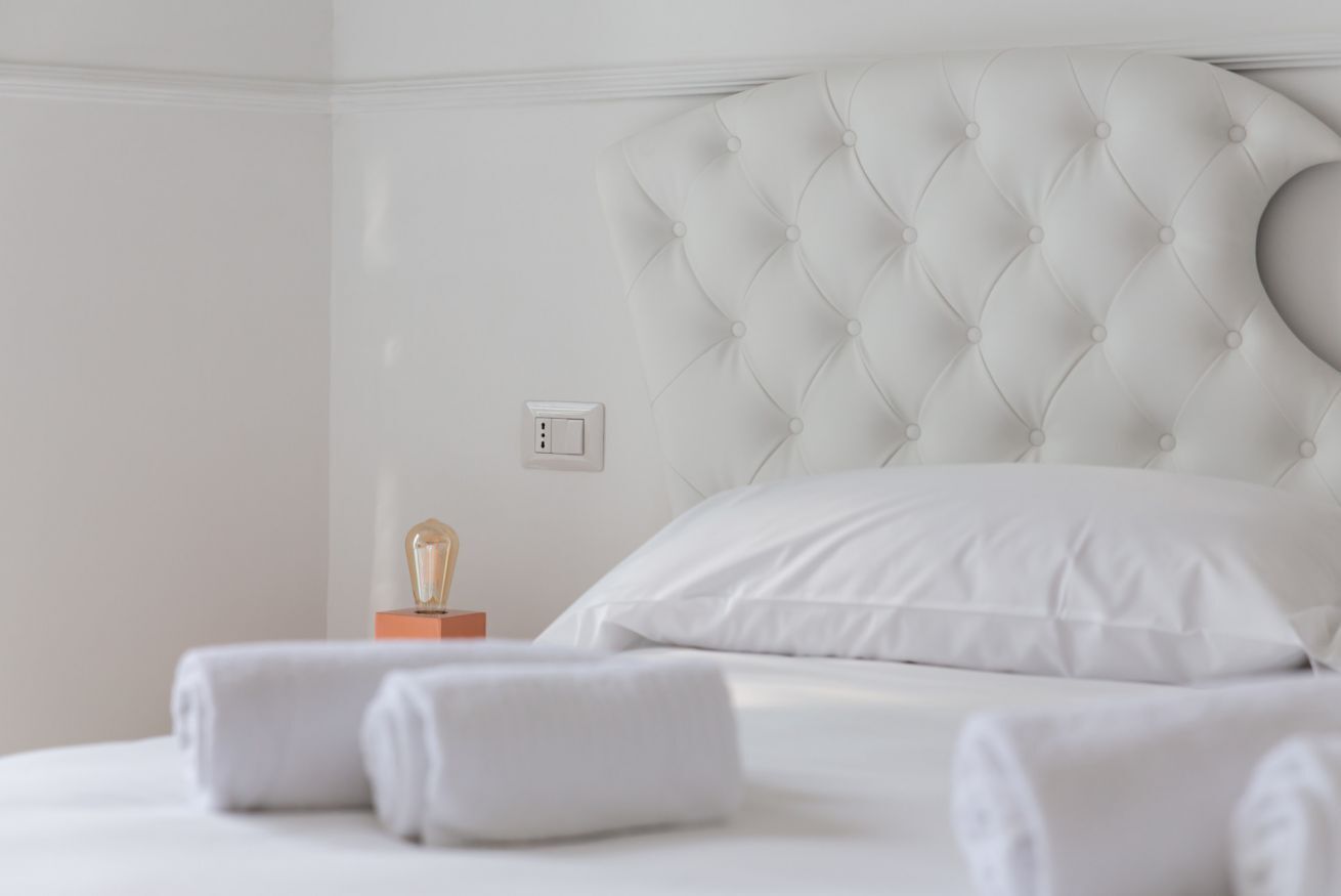 A neatly made bed with white linens and a tufted headboard, a bedside table with a lamp, and a wall socket.